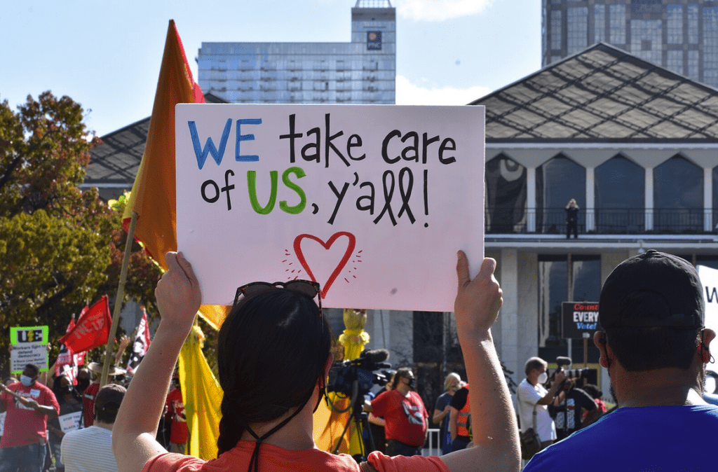 A person in a small crowd holds up a sign that says “WE take care of US, ya’ll!”